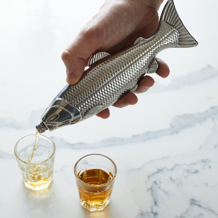 Hand holding the Fish Flask and pouring liquid into shot glasses on a counter top
