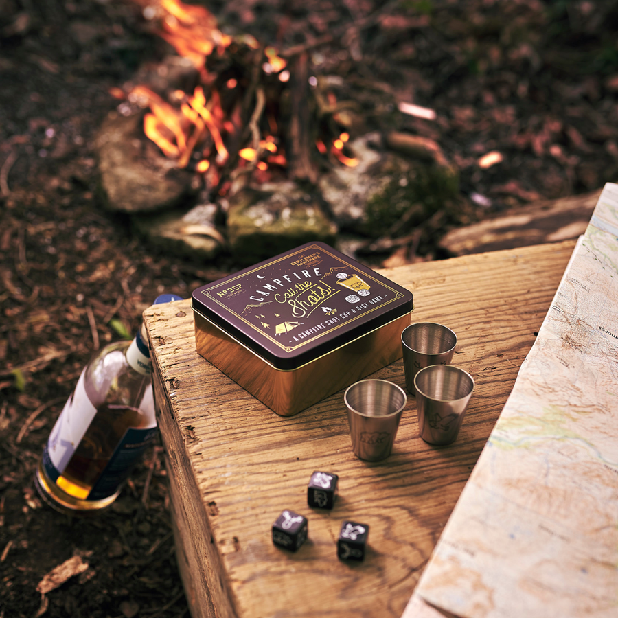 Campfire Call The Shots Game with metal tin, 3 shot glasses, and 3 dice in front of campfire