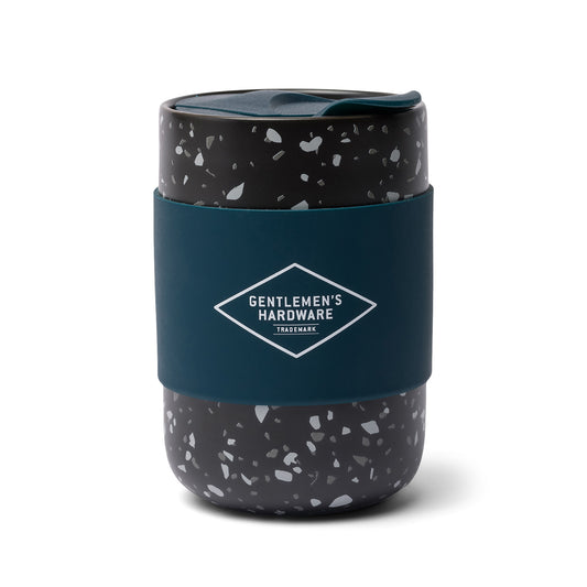 The Ceramic Travel Coffee Mug 13.5 oz features a black and white speckled design, a dark teal silicone sleeve, and Gentlemens Hardware branding—combining style and practicality for drinks on the go.