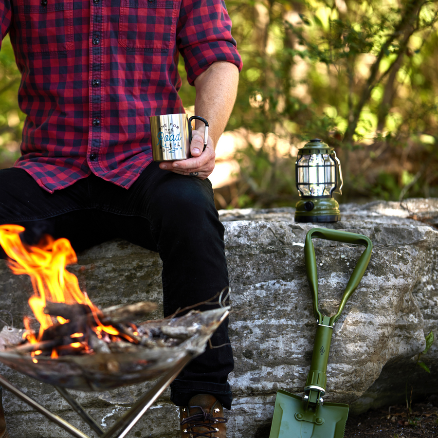 Folding Shovel leaning against a log holding a human with a mug and lantern; fire pit in the foreground